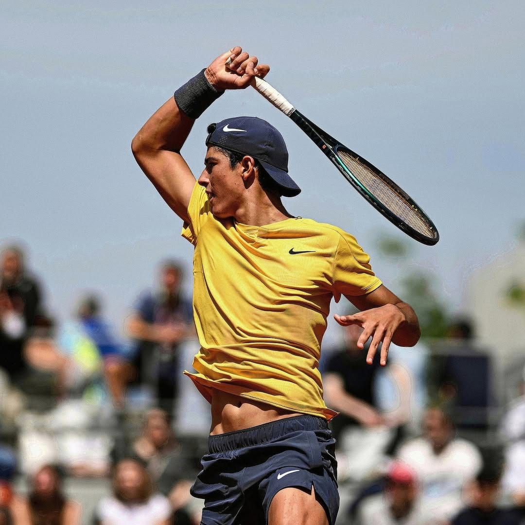 Federico Cinà follows through on a forehand swing in yellow Nike kit and navy cap, crowd blurred behind him.