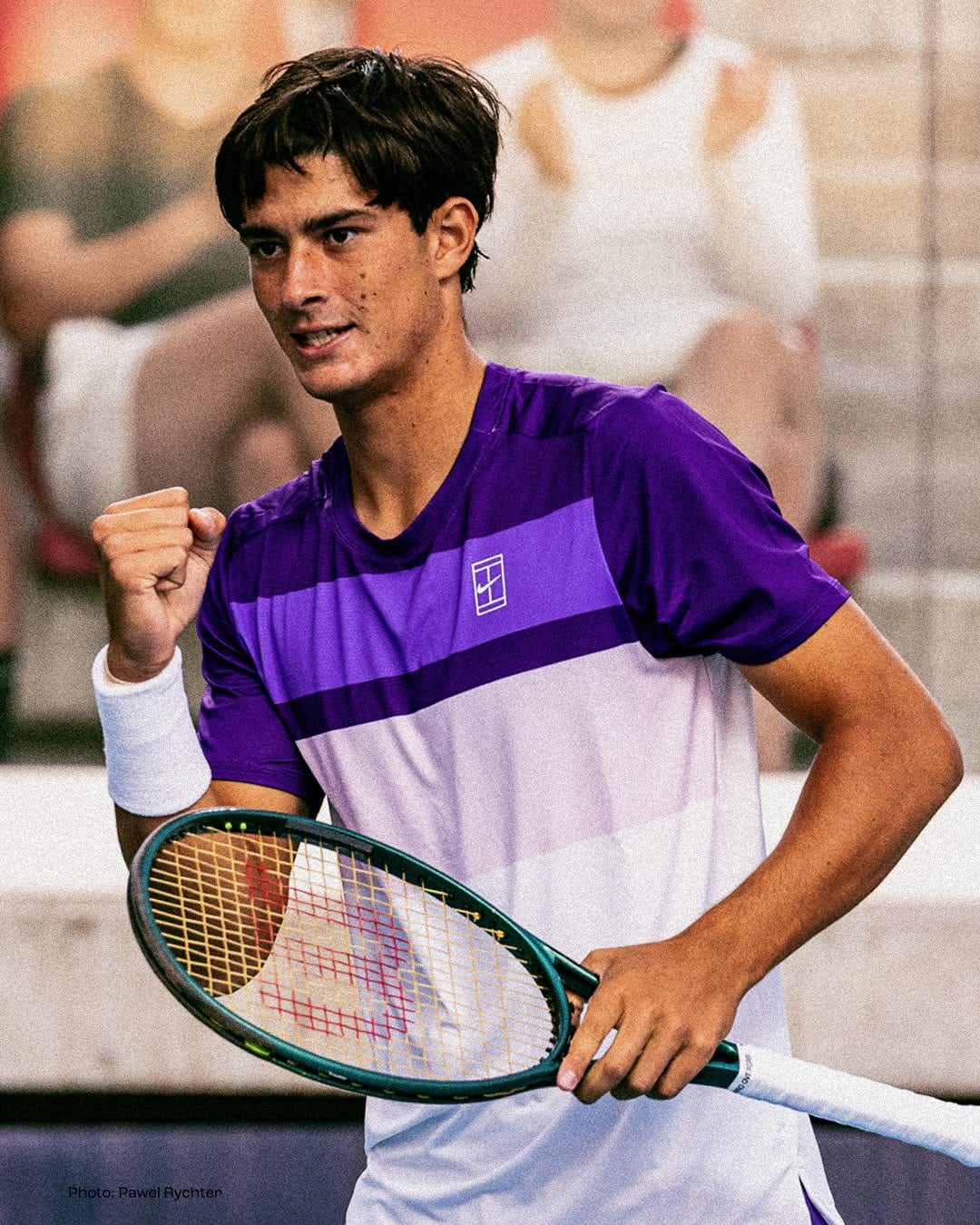 Federico Cinà pumps his fist in celebration wearing a purple Nike shirt, holding his racket at an outdoor hard court.