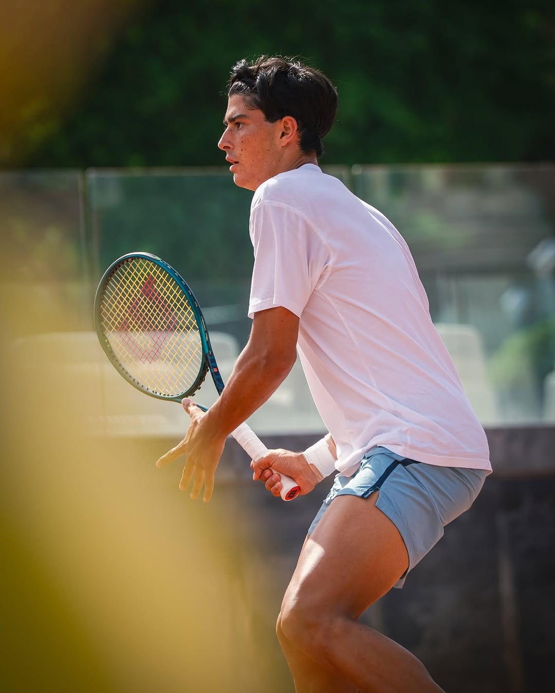 Federico Cinà crouches in ready position holding a Wilson racket, wearing white tee and light blue shorts, in bright outdoor sunlight.