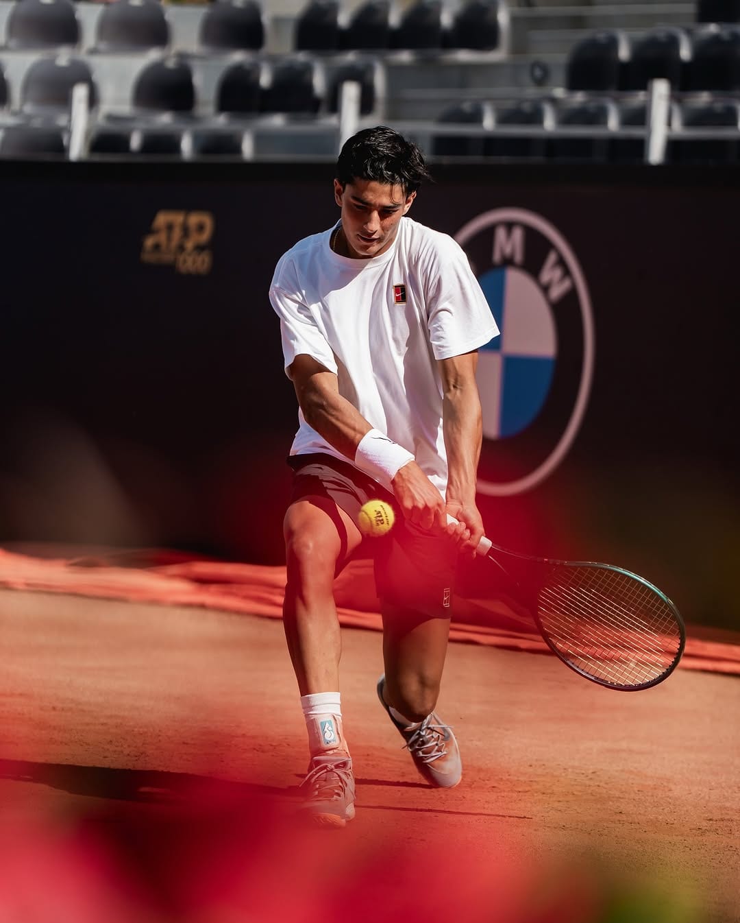 Federico Cinà plays a low backhand on clay at an ATP 1000 event, BMW and ATP logos visible on the hoarding behind him.