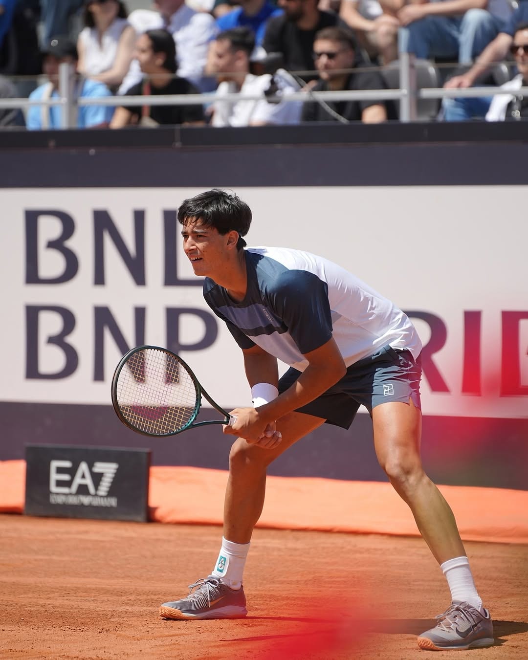 Federico Cinà crouches in a ready position on clay at the Internazionali BNL d'Italia, racket held low, crowd behind him, EA7 and BNP banners visible.