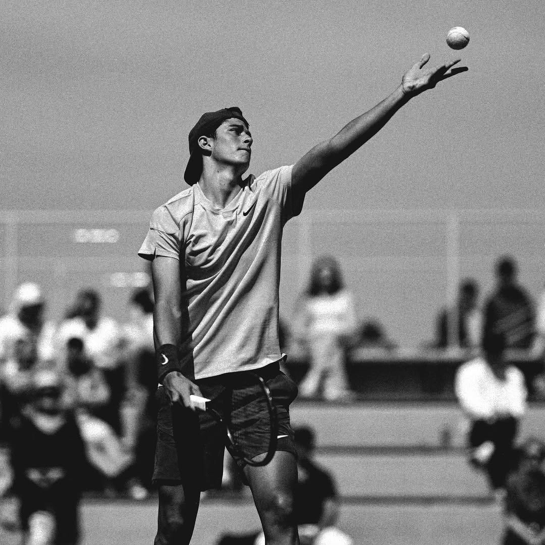 Federico Cinà tosses the ball upward mid-serve, wearing a Nike shirt and cap, with a blurred crowd behind him, black-and-white.