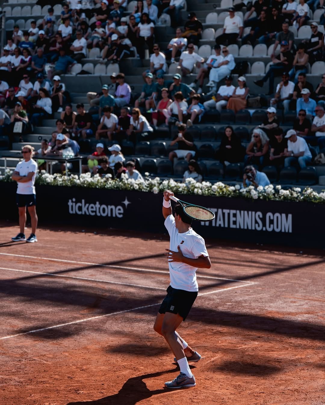 Federico follows through on a shot on red clay, racket raised, with Ticketone and WTATennis.com courtside boards and a packed grandstand behind.