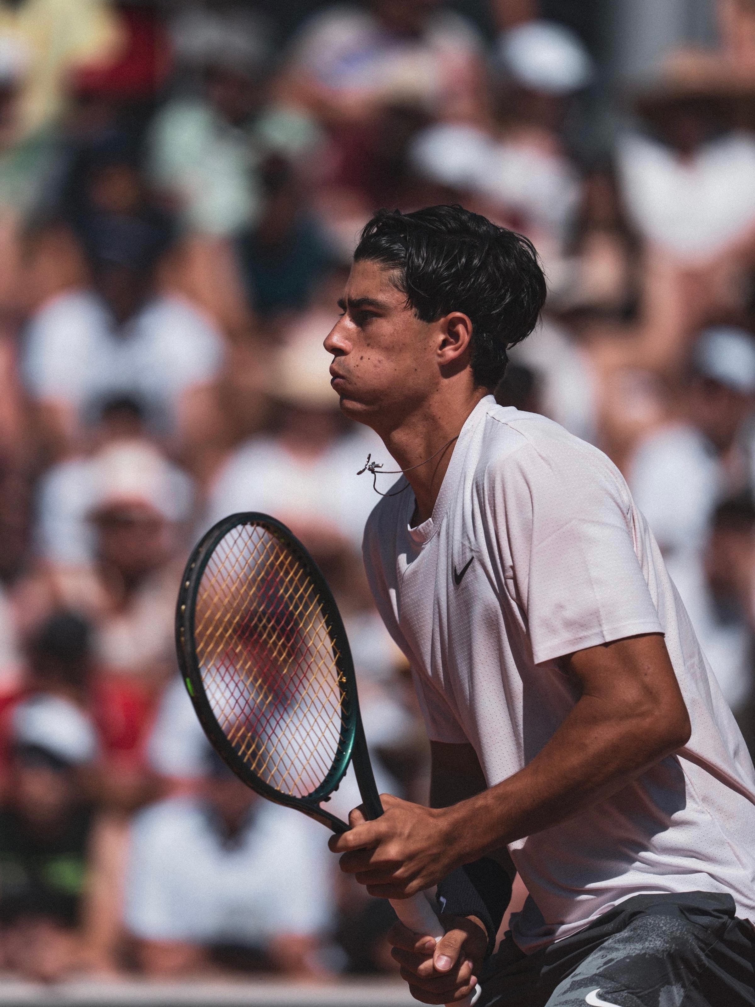 Federico Cinà holds his racket in ready position, focused expression, crowd blurred in bright sunlight behind him.