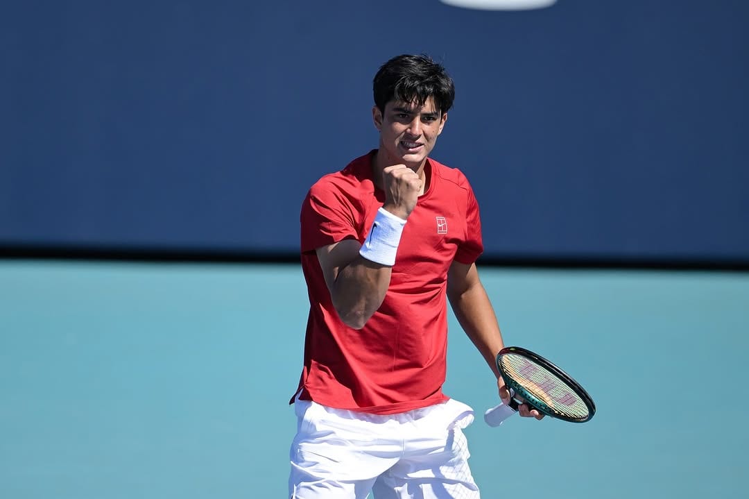Federico Cinà pumps his fist in celebration on a hard court, wearing a red Nike shirt and white wristband, racket in hand.