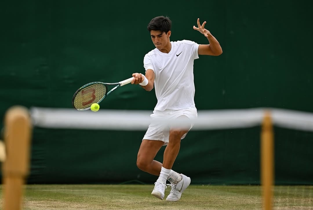 Federico Cinà hits a low forehand on a Wimbledon grass court, in all-white Nike kit, mid-follow-through.
