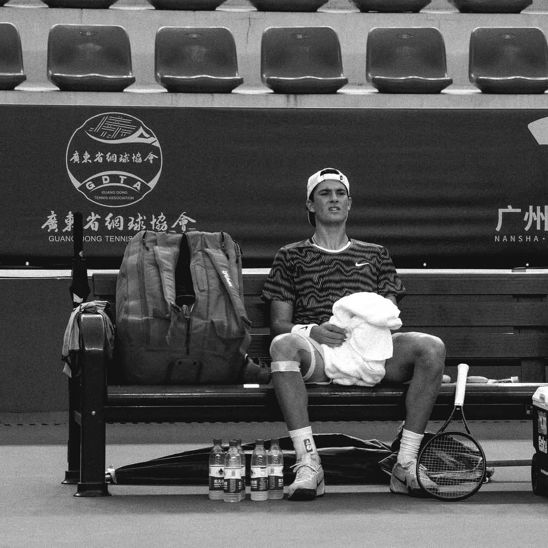 Federico sits on a courtside bench holding a towel, racket at his feet, Guang Dong Tennis Association branding visible behind him.