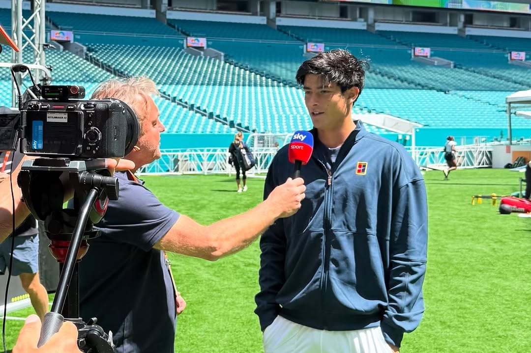 Federico Cinà speaks into a Sky Sport microphone during a TV interview on a grass area inside a large open-air stadium, wearing a Nike Court jacket.