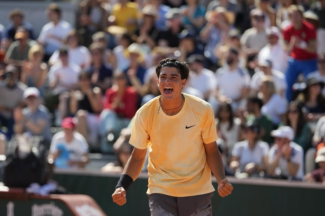 Federico Cinà roars and pumps both fists in celebration on clay, wearing a yellow Nike shirt, packed crowd behind him.
