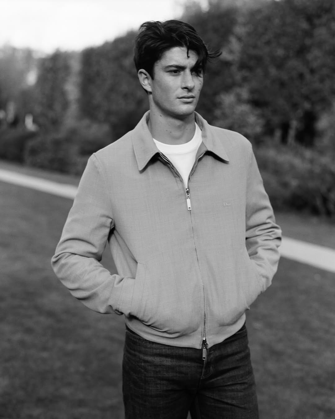 Federico Cinà stands outdoors in a Dior zip jacket and jeans, hands in pockets, gazing off-camera, trees in background.