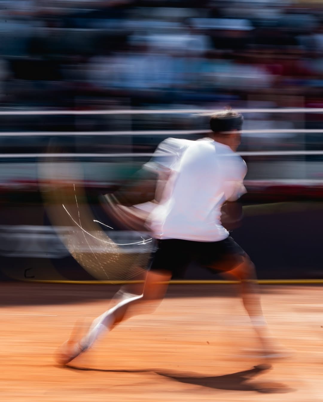 Federico sprints on red clay in a motion-blurred shot, white shirt and headband, racket swinging, packed stands behind him.