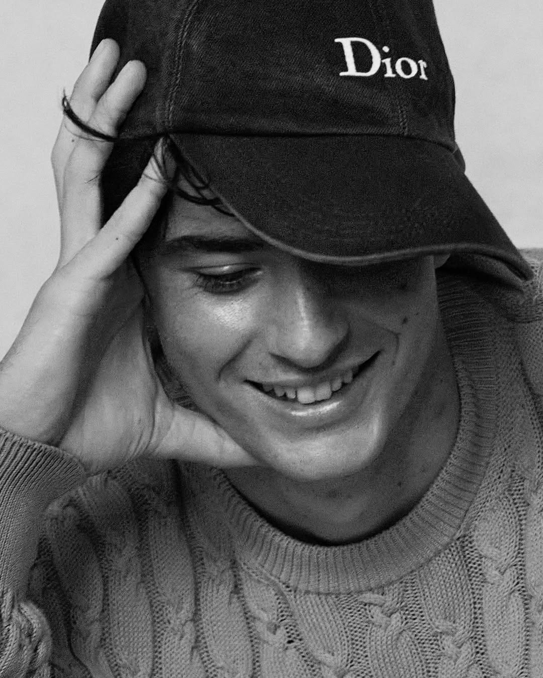 Federico Cinà smiles downward, hand on a Dior cap, wearing a cable-knit sweater, black-and-white studio portrait.