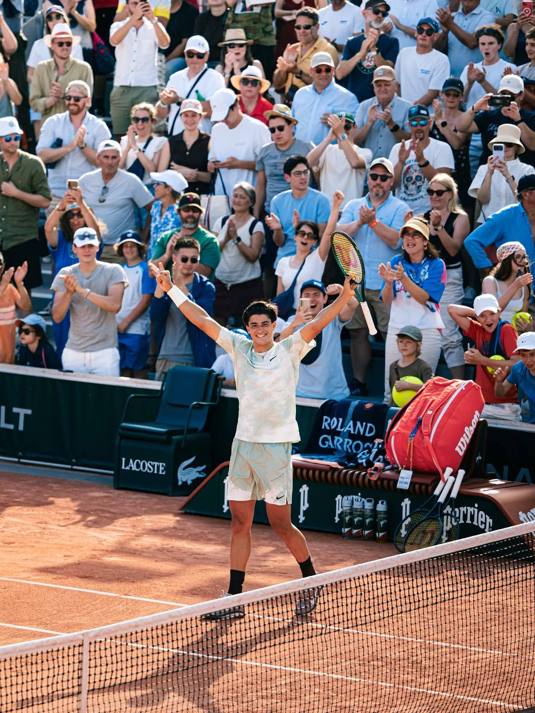 Federico Cinà raises both arms in celebration on red clay at Roland Garros, crowd applauding behind him, Lacoste and Perrier boards visible.