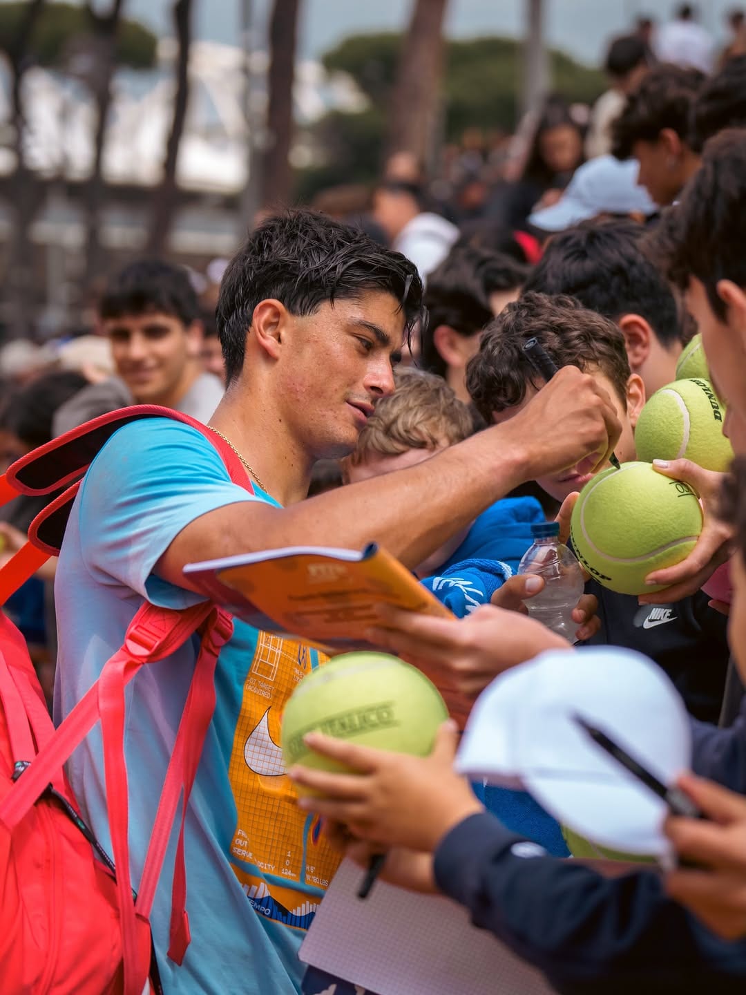Federico Cinà signs autographs for a crowd of fans, holding Dunlop tennis balls and a notebook, red backpack over his shoulder.